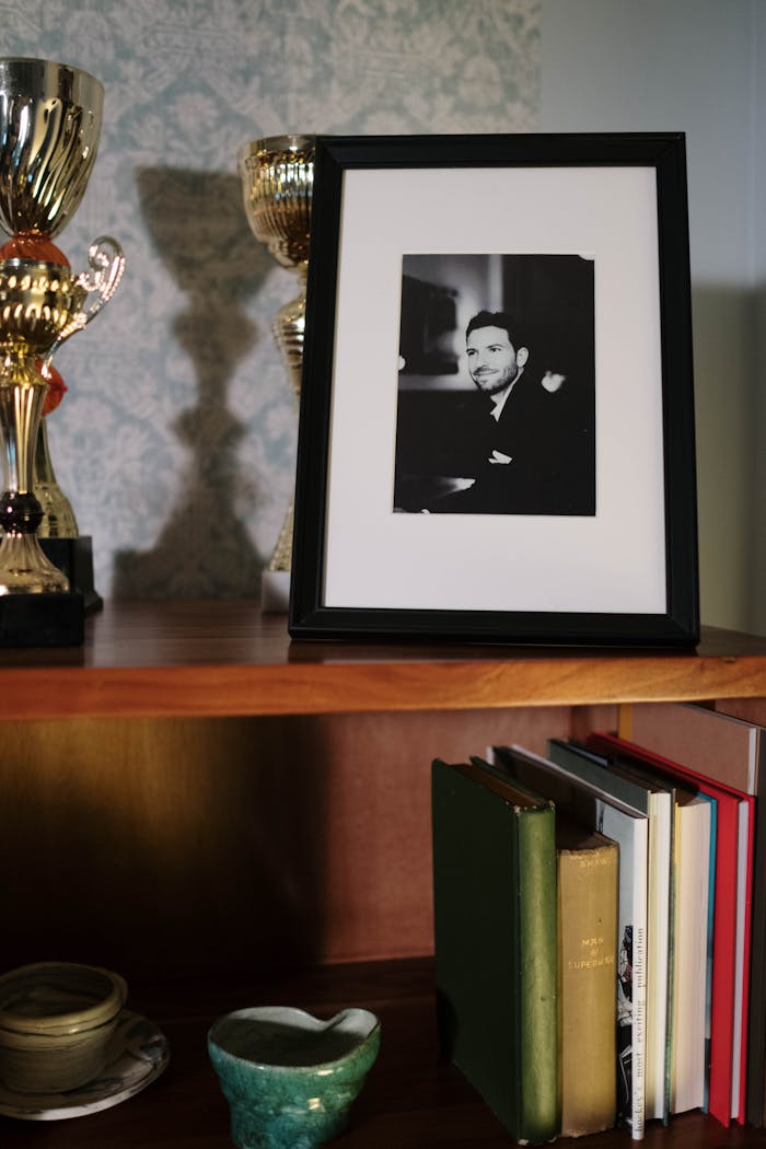 about-us A framed black-and-white portrait alongside trophies on a wooden shelf with books.